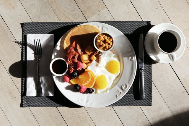 white plate on a black table mat with brunch items including sunny side up eggs, bacon, potato pieces, pancakes with syrup, and a fruit medley next to a cup of coffee